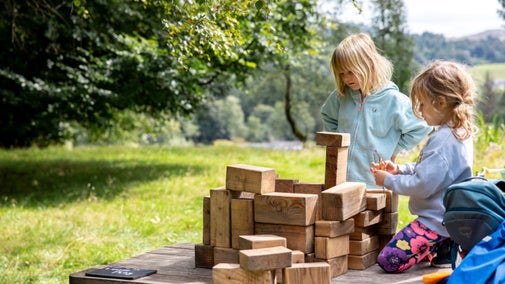 Children building a large stack of wooden blocks on a table in a grassy garden at Allan Bank, with trees and countryside in the background.